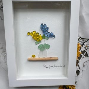 May include: A framed piece of art featuring a flower arrangement made of blue and yellow sea glass. The piece is titled "The Farmhouse Floret".