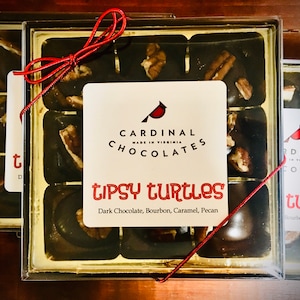 May include: A box of Cardinal Chocolates Tipsy Turtles, a dark chocolate, bourbon, caramel, and pecan treat. The box is tied with a red ribbon and has a white label with the company logo and product name.