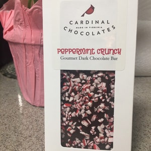 May include: A white box with a red and white logo that says "Cardinals Chocolates Made in Virginia". The box contains a dark chocolate bar with peppermint pieces. The bar is labeled "Peppermint Crunch Gourmet Dark Chocolate Bar".
