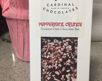 May include: A white box with a red and white logo that says "Cardinals Chocolates Made in Virginia". The box contains a dark chocolate bar with peppermint pieces. The bar is labeled "Peppermint Crunch Gourmet Dark Chocolate Bar".