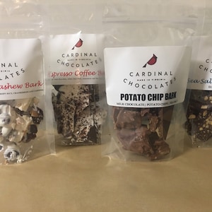 May include: Four bags of Cardinal Chocolates, each with a different flavor: Cranberry Cashew Bark, Espresso Coffee Bark, Potato Chip Bark, and Sea Salt Almond Bark. The bags are clear plastic with white labels featuring a red cardinal logo.