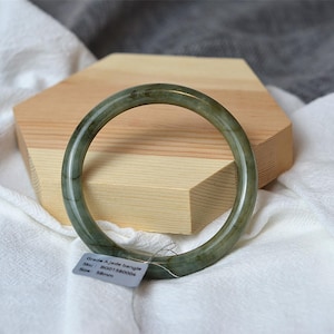 Può includere: Un braccialetto di giada verde, appoggiato su un blocco di legno e tessuto bianco. Il braccialetto circolare ha una superficie liscia e un'etichetta con dettagli del prodotto. Il diametro del braccialetto è di 5,8 cm.