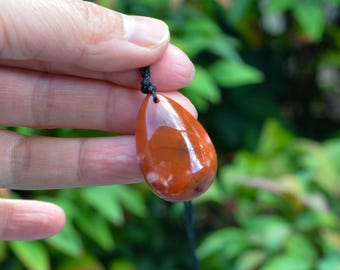May include: A teardrop-shaped pendant with a glossy, reddish-brown surface, suspended from a black cord. The pendant has a polished appearance with lighter, white-toned inclusions. The background is blurred green foliage.