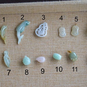 May include: A selection of twelve jade pendants in diverse shapes and colours, including a dolphin, a butterfly, and abstract forms. The pendants are displayed on a beige fabric surface.