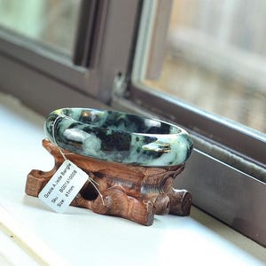 Könnte beinhalten: Ein Jade-Armreif mit einem dunkelgrünen und schwarzen Muster, präsentiert auf einem geschnitzten Holzständer. Der Armreif hat eine Größe von etwa 6,1 cm. Ein Etikett am Ständer trägt die Aufschrift "Grace A. Jade Bangle". Der Hintergrund umfasst ein Fenster und natürliches Licht.