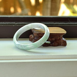 May include: A pale green jade bangle bracelet. The smooth, polished bracelet is resting on a small, carved wooden stand. The jade has a translucent quality, with subtle variations in color.