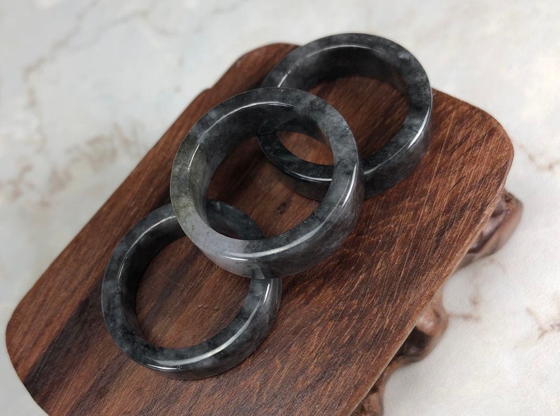 May include: Three black jade bangle bracelets stacked on a wooden stand. The bracelets are smooth and polished.