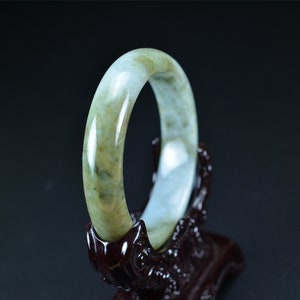 May include: A jade bangle bracelet with a light green and white color scheme. The bangle is smooth and polished, and it is displayed on a wooden stand.