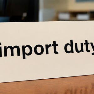 May include: A rectangular, off-white sign with the words "import duty" printed in bold, black letters. The sign is placed on a wooden surface, with a blurred background.