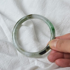 May include: A round jade bangle with shades of green, white, and black. The smooth, polished surface of the bracelet is held against a white fabric background. The jade is a natural stone.