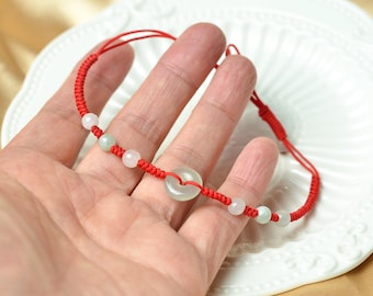 May include: Red string bracelet with white and green beads and a central jade disc. The bracelet is displayed on a white plate. The bracelet is a delicate piece of jewelry.