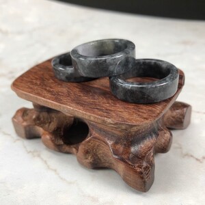 May include: Three gray jade rings stacked on a wooden stand. The rings are smooth and have a mottled pattern.