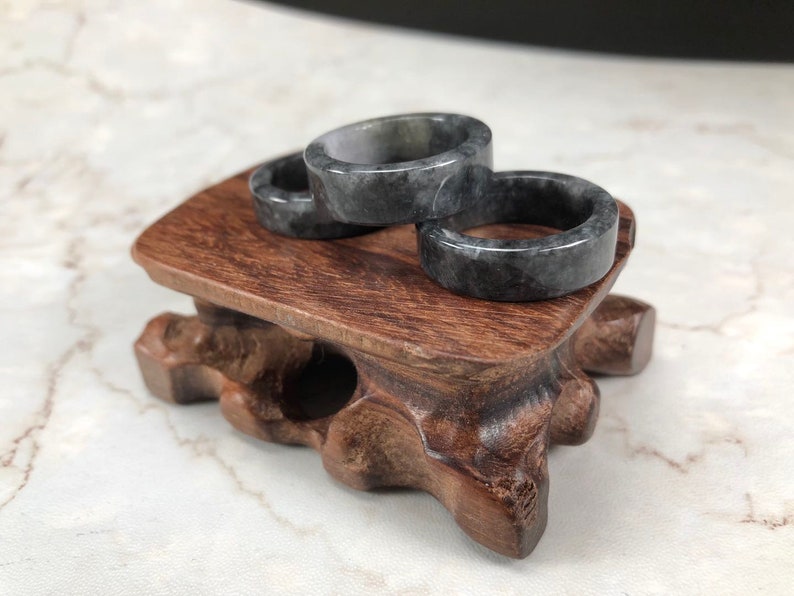 May include: Three gray jade rings stacked on a wooden stand. The rings are smooth and have a mottled pattern.