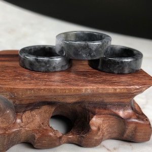 May include: Three dark gray jade rings stacked on a wooden stand. The rings are smooth and polished, with a subtle marbling effect.