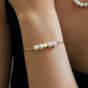 May include: A gold necklace and bracelet set featuring white pearls. The necklace has a delicate chain with five pearls. The bracelet has a similar design with five pearls and a gold heart charm with the letter "S".