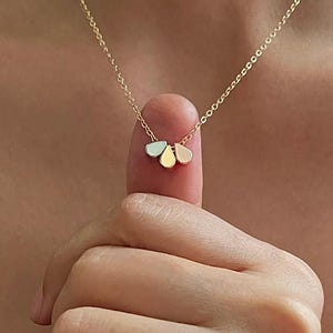 Peut inclure: Un délicat collier en or avec un pendentif composé de trois petits charmes en forme de pétales aux couleurs pastel. Le collier est tenu par un doigt, mettant en valeur la fine chaîne et les teintes subtiles des charmes. C'est un bijou.