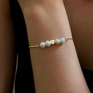 May include: A gold bracelet and necklace set featuring white pearls. The bracelet has a heart-shaped charm with the letter "S". The necklace has a delicate chain with a series of pearls. The jewelry is displayed on a person's arm and chest.