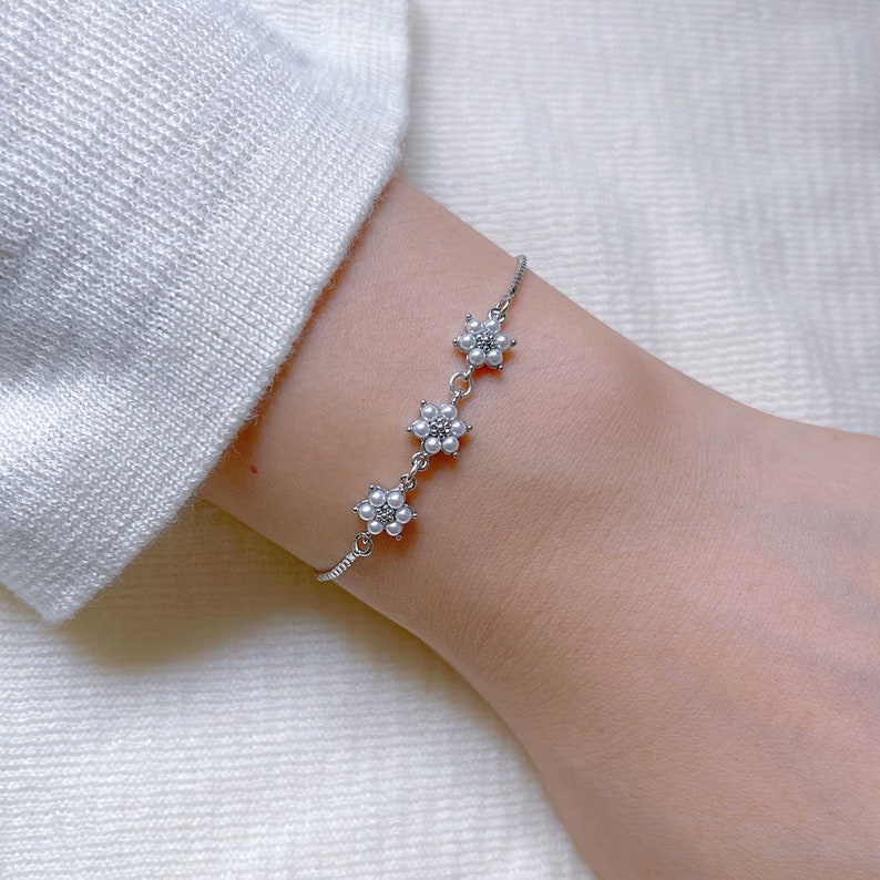 May include: A silver bracelet with three flower charms made of white pearls. The bracelet is on a person's wrist.