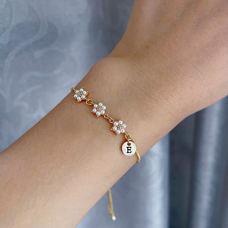 K&ouml;nnte beinhalten: Ein goldenes Kettenarmband mit einem Anh&auml;nger, auf dem "E" steht, und drei Blumenanh&auml;ngern mit wei&szlig;en Perlen.