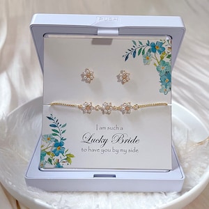 May include: A white gift box with a gold bracelet and earrings. The box has a floral design and the text "I am such a Lucky Bride to have you by my side".
