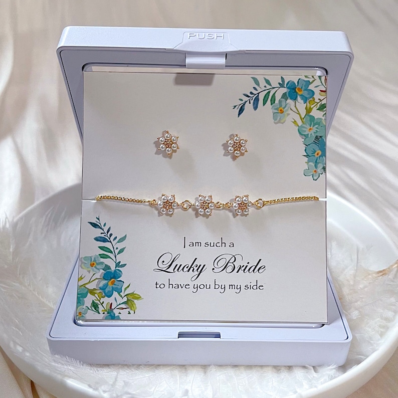 May include: A white gift box with a gold bracelet and earrings. The box has a floral design and the text "I am such a Lucky Bride to have you by my side".