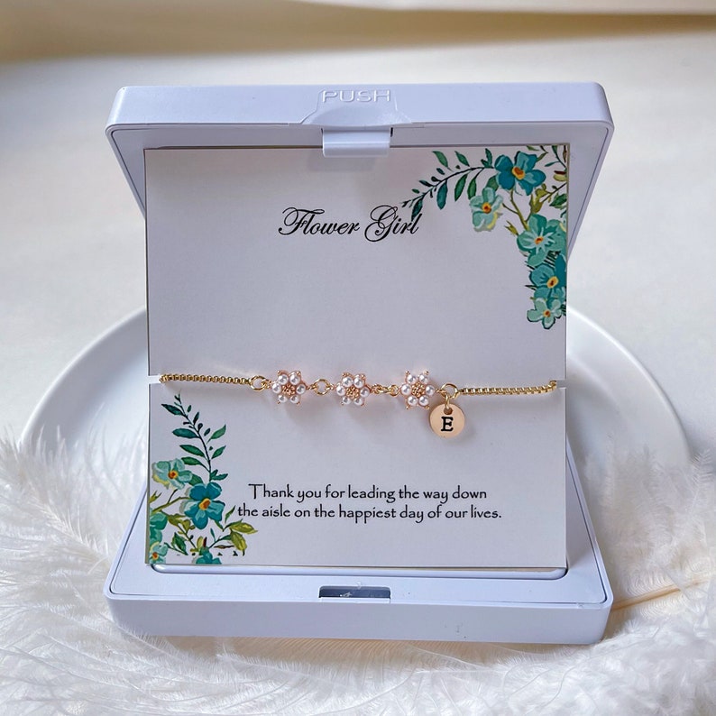 K&ouml;nnte beinhalten: Ein goldenes Kettenarmband mit drei Blumenanh&auml;ngern und einem Buchstaben "E" Anh&auml;nger. Das Armband befindet sich in einer wei&szlig;en Schachtel mit einer Karte, auf der "Flower Girl" und "Vielen Dank, dass du den Weg zum Altar am gl&uuml;cklichsten Tag unseres Lebens gef&uuml;hrt hast." steht.