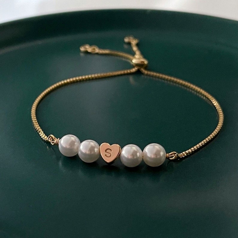 Puede incluir: Un brazalete de cadena dorada con un dije en forma de coraz&oacute;n y cinco perlas blancas. El dije en forma de coraz&oacute;n tiene la letra "S" grabada.