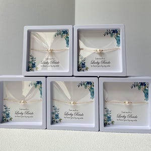 May include: Five white boxes with blue floral designs and clear lids containing delicate gold chain bracelets with small pearl accents. Each box has a white label with the text "I am such a Lucky Bride to have you by my side."