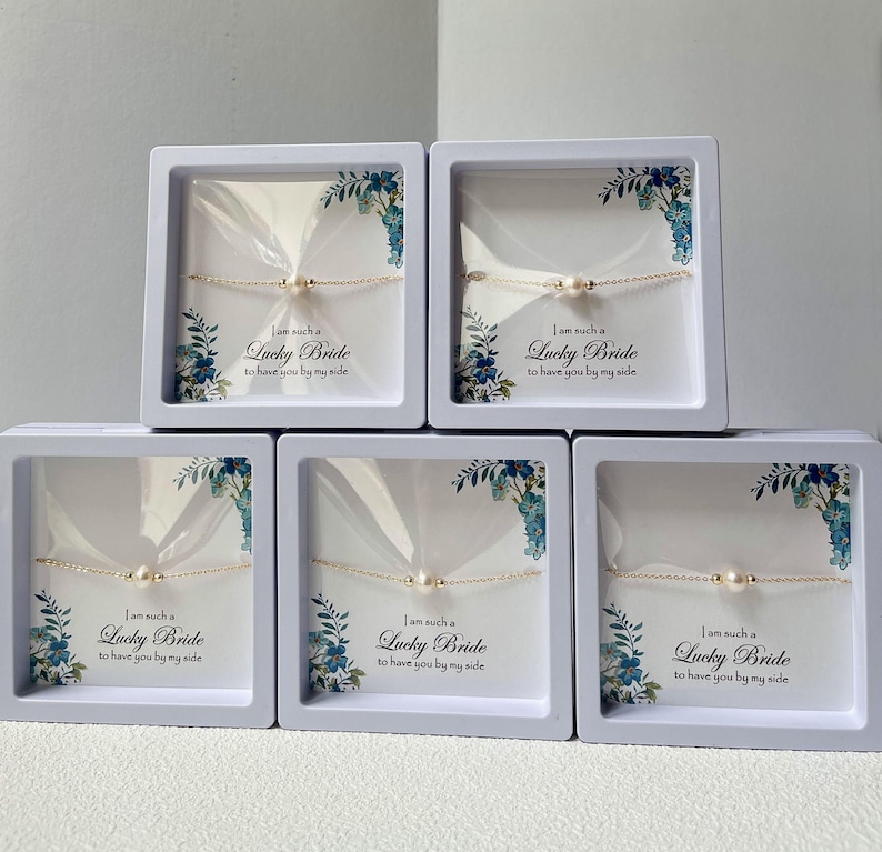 May include: Five white boxes with blue floral designs and clear lids containing delicate gold chain bracelets with small pearl accents. Each box has a white label with the text "I am such a Lucky Bride to have you by my side."