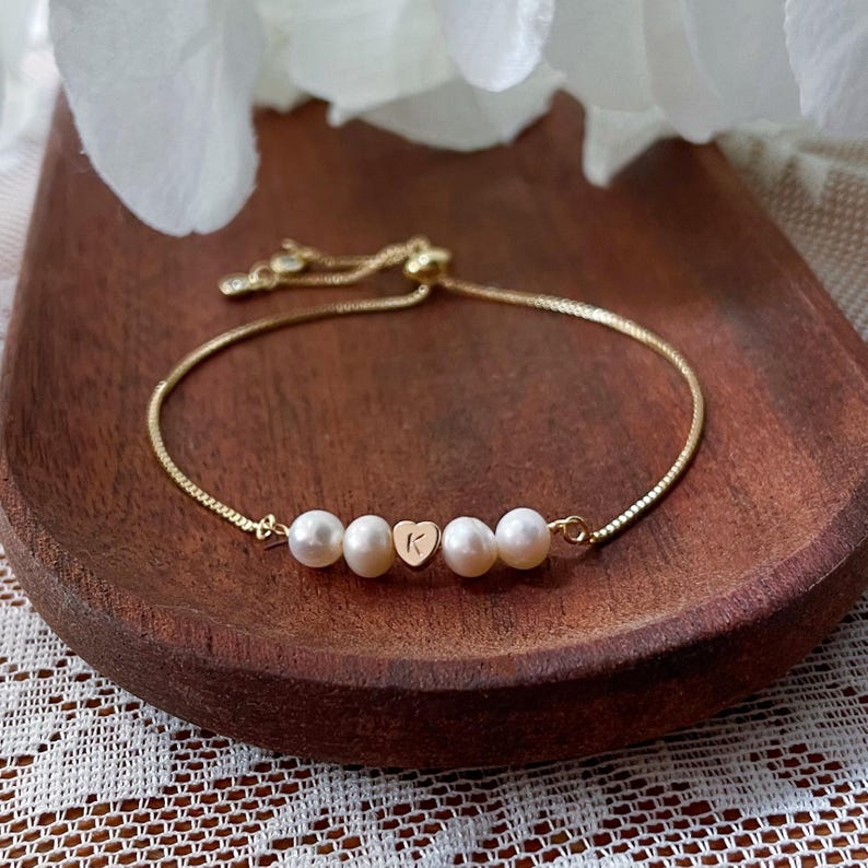 May include: Gold-toned bracelet featuring four white pearls and a heart-shaped charm with the letter "K". The adjustable bracelet is displayed on a wooden surface, showcasing its delicate design.