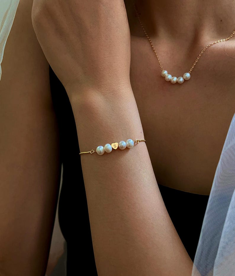 May include: A gold bracelet and necklace set featuring white pearls. The bracelet has a heart-shaped charm with the letter "S". The necklace has a delicate chain with a series of pearls. The jewelry is displayed on a person's arm and chest.