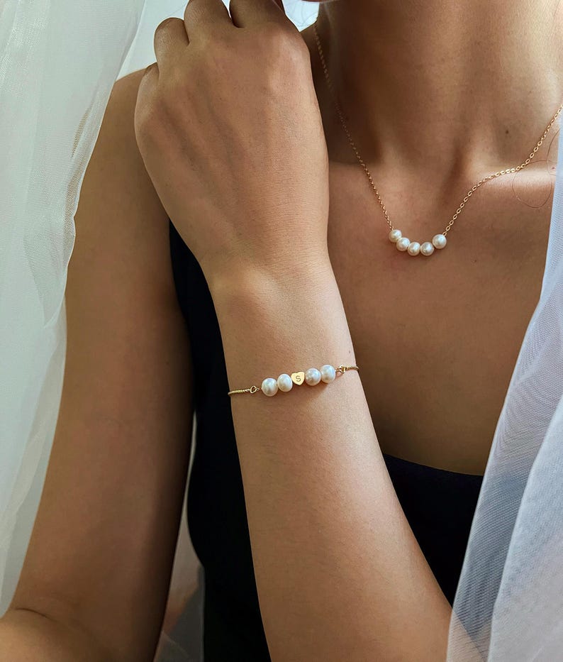 May include: A gold necklace and bracelet set featuring white pearls. The necklace has a delicate chain with five pearls. The bracelet has a similar design with five pearls and a gold heart charm with the letter "S".