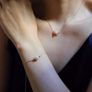 May include: A delicate gold chain necklace and bracelet with a single pearl pendant on each. The necklace and bracelet are worn by a person with fair skin.