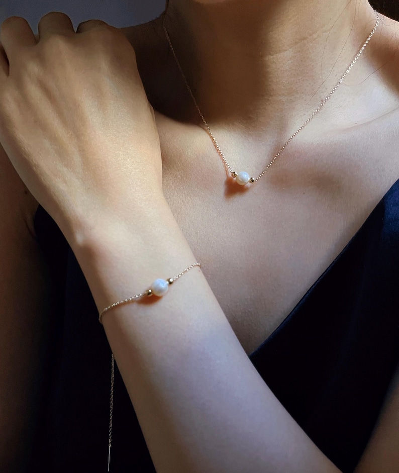 May include: A delicate gold chain necklace and bracelet with a single pearl pendant on each. The necklace and bracelet are worn by a person with fair skin.