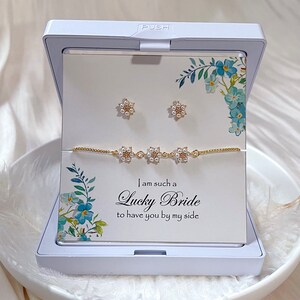 K&ouml;nnte beinhalten: Eine wei&szlig;e Geschenkbox mit einem goldenen Armband und Ohrringen. Die Box hat ein Blumenmuster und den Text "Ich bin so eine gl&uuml;ckliche Braut, dich an meiner Seite zu haben".
