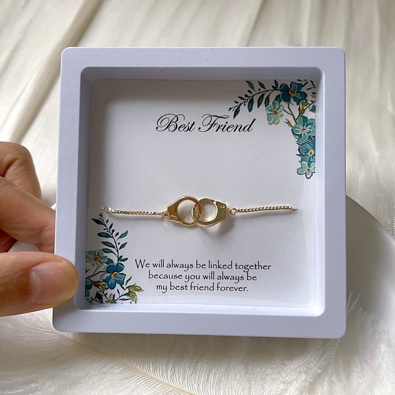 K&ouml;nnte beinhalten: Eine wei&szlig;e Box mit einem goldenen Armband mit zwei verbundenen Ringen. Die Box enth&auml;lt eine wei&szlig;e Karte mit dem Text "Best Friend" und einer Nachricht, die lautet: "Wir werden immer miteinander verbunden sein, weil du f&uuml;r immer mein bester Freund sein wirst."