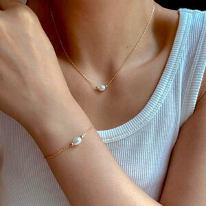 May include: A delicate gold chain necklace and bracelet with a single pearl pendant on each. The necklace and bracelet are worn by a person.