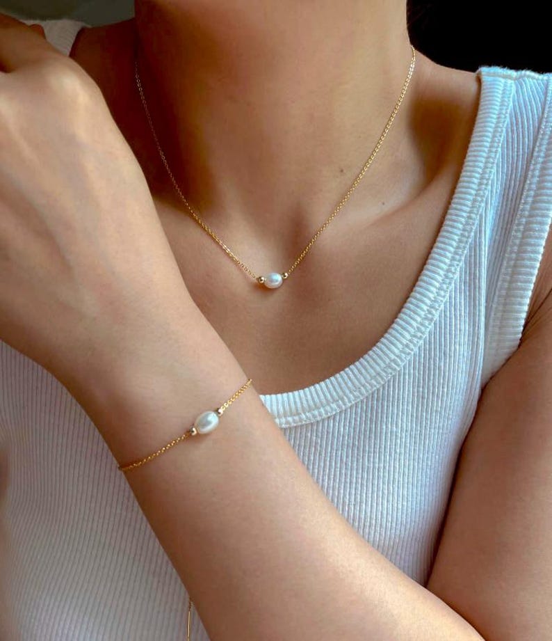 May include: A delicate gold chain necklace and bracelet with a single pearl pendant on each. The necklace and bracelet are worn by a person.