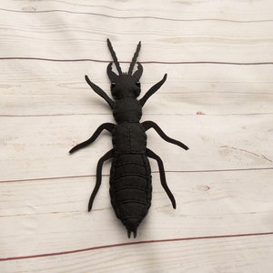 May include: A black felt insect with long legs and a segmented body. The insect is lying on a white wooden surface.