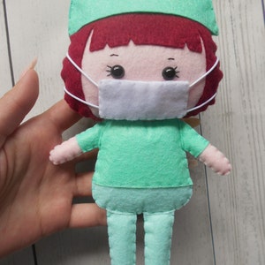 May include: A felt doll wearing a green scrub top, pants, and a surgical cap. The doll has red hair, a white face mask, and black eyes.