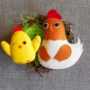 K&ouml;nnte beinhalten: Zwei Filz-Spielzeug-H&uuml;hner in einem Nest. Das gr&ouml;&szlig;ere Huhn ist braun mit wei&szlig;en Akzenten und hat einen roten Kamm und einen roten Kehllappen. Das kleinere Huhn ist gelb mit einem roten Kamm und schwarzen Augen.