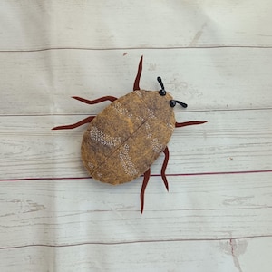 May include: A brown and white beetle with red legs and black antennae. The beetle is made of felt and is a decorative item.