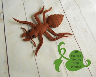 May include: A brown felt ant with black eyes. The ant is lying on a light wood surface. A green leaf with the text "PDF pattern (step by step photos)" is in the lower right corner of the image.