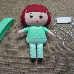 May include: A felt doll with red hair, a green shirt, and green pants. The doll is holding a white felt mask with white string ties.