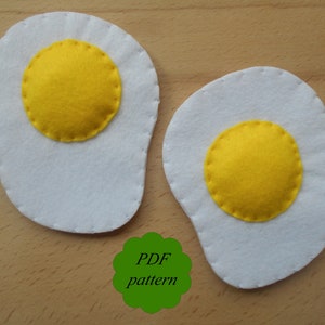 May include: Two felt egg shapes with white egg whites and yellow yolks. The yolks are circular and stitched onto the white egg shapes. The image also includes a green scalloped shape with the text "PDF pattern".