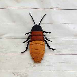 May include: A brown and black felt cockroach with segmented legs and a black head. The cockroach is on a white wooden background.