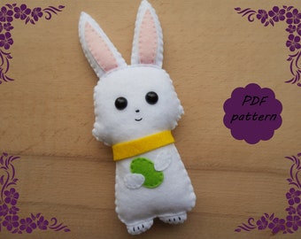 May include: A white felt bunny holding a green felt egg. The bunny has pink ears, black eyes, and a yellow felt collar. The bunny is on a wooden surface with a purple floral border.