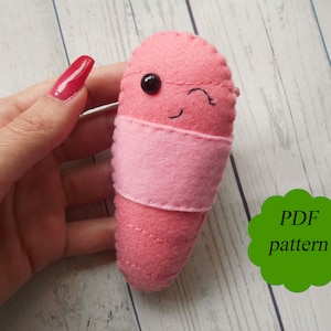 May include: A pink felt worm with a black eye and a stitched-on smile. The worm is wearing a pink felt band around its middle. The text "PDF pattern" is written on a green circle.