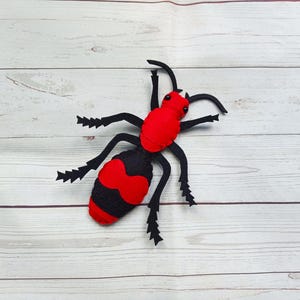 May include: A handcrafted felt insect toy featuring a vibrant red head and thorax with a black and red striped abdomen. The creature has six jagged black legs and two long antennae, with small black bead eyes positioned on the red head section.