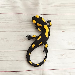 Puede incluir: Un juguete de salamandra hecho a mano de fieltro negro con manchas amarillas. La salamandra tiene una cola curvada y pequeños pies negros. Colocado sobre una superficie de madera clara.